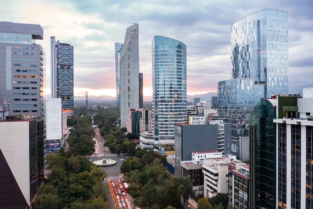 Rascacielos modernos a lo largo del Paseo de la Reforma en la Ciudad de México al atardecer, con espacios verdes abajo y montañas en la distancia.
