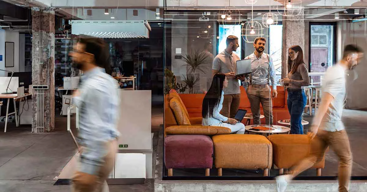 Modern open office with glass partitions, colorful furniture, and people working and moving in various areas.