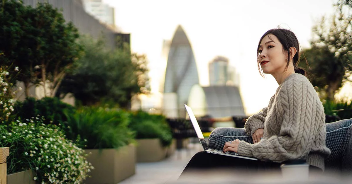 A woman sitting on a park bench, focused on her laptop, with trees and a pathway in the background.