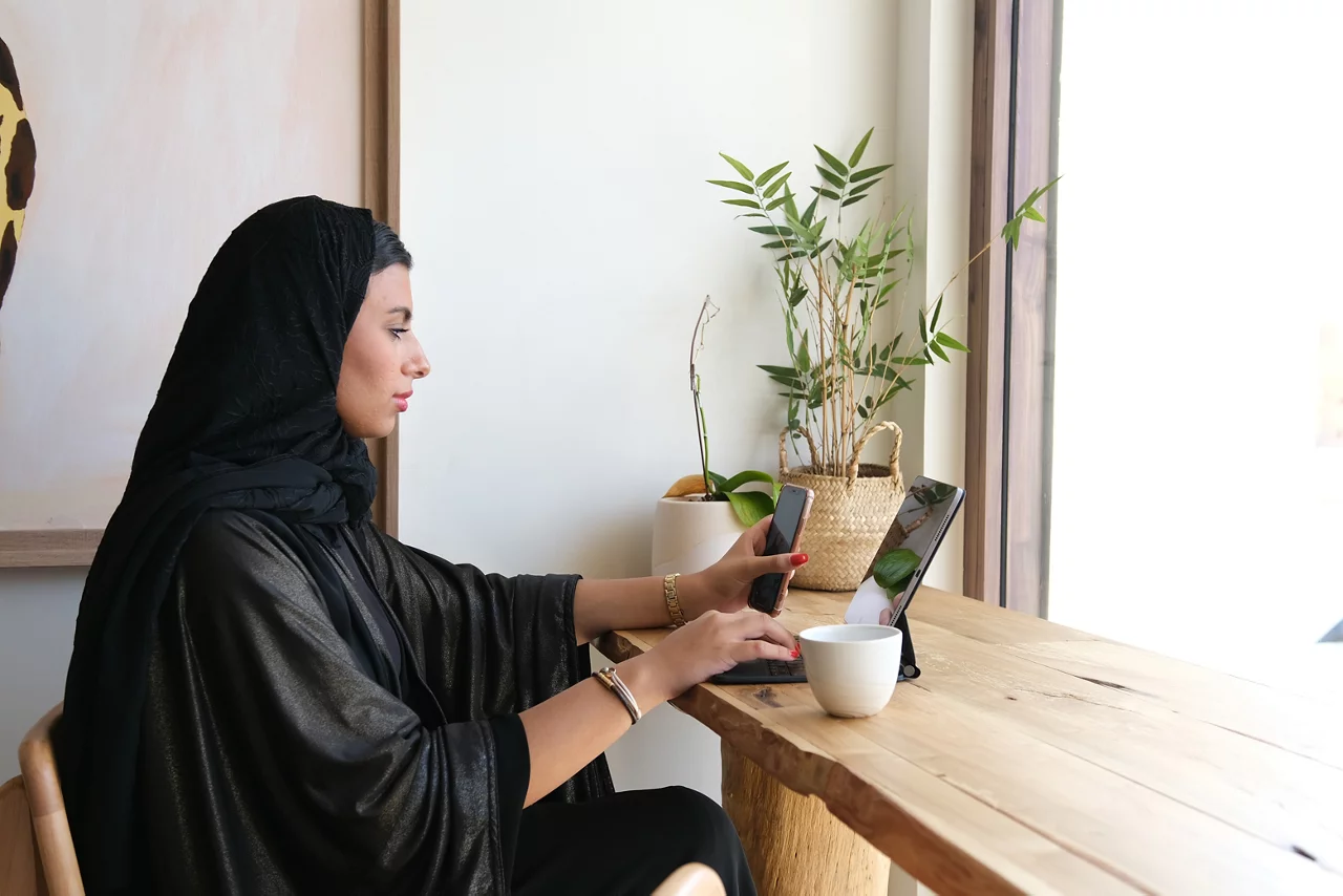 Professional woman in black hijab working on tablet at wooden desk with coffee and plants in modern office space.