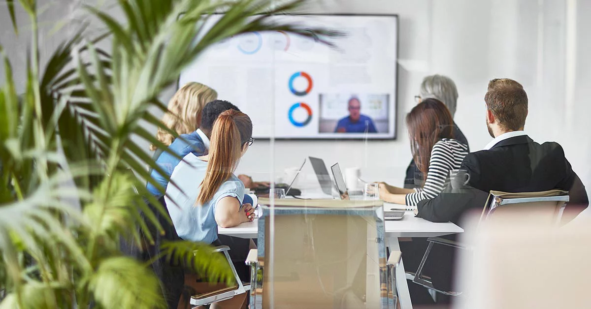 A group of people seated in a meeting room, focused on a large screen displaying a presentation.