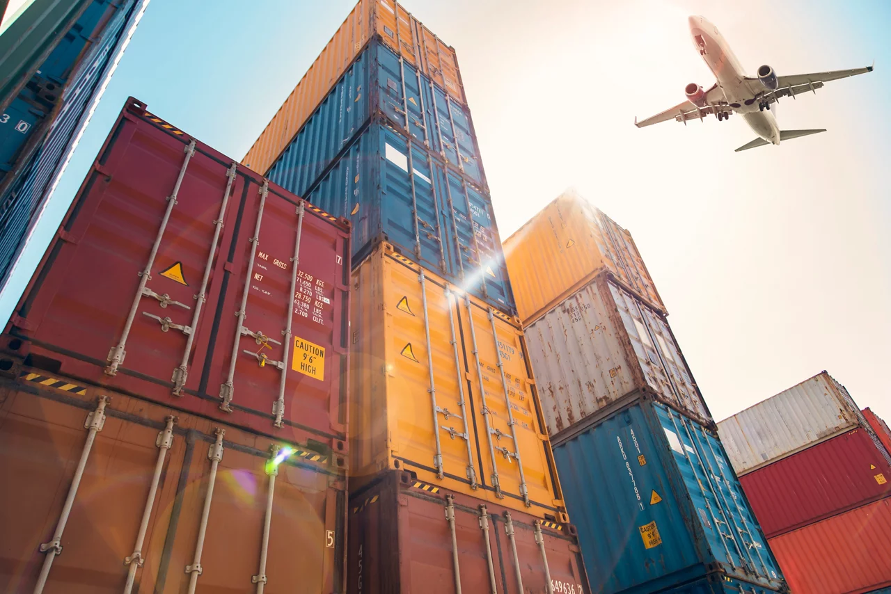 Stacked shipping containers at a port with airplane flying overhead against bright sky, representing global logistics.
