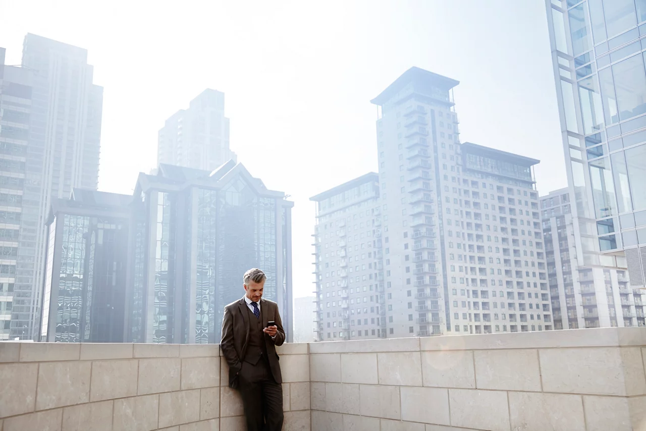 Businessman using smartphone on urban rooftop, surrounded by skyscrapers in a city skyline.