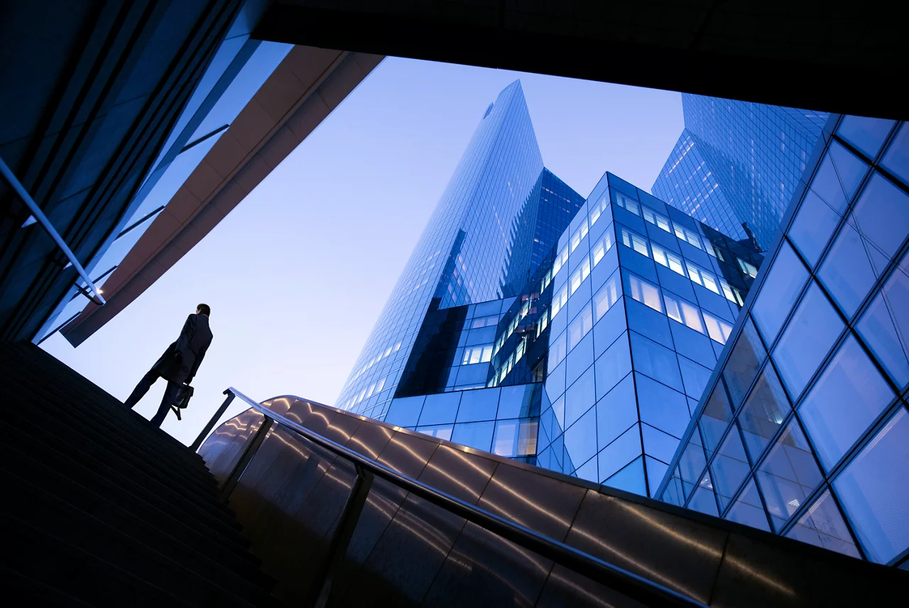 Business professional ascending stairs toward modern glass skyscrapers in downtown financial district at dusk.