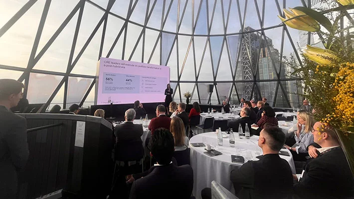Business meeting in modern glass-walled venue with city view. Presenter addressing audience at round tables.