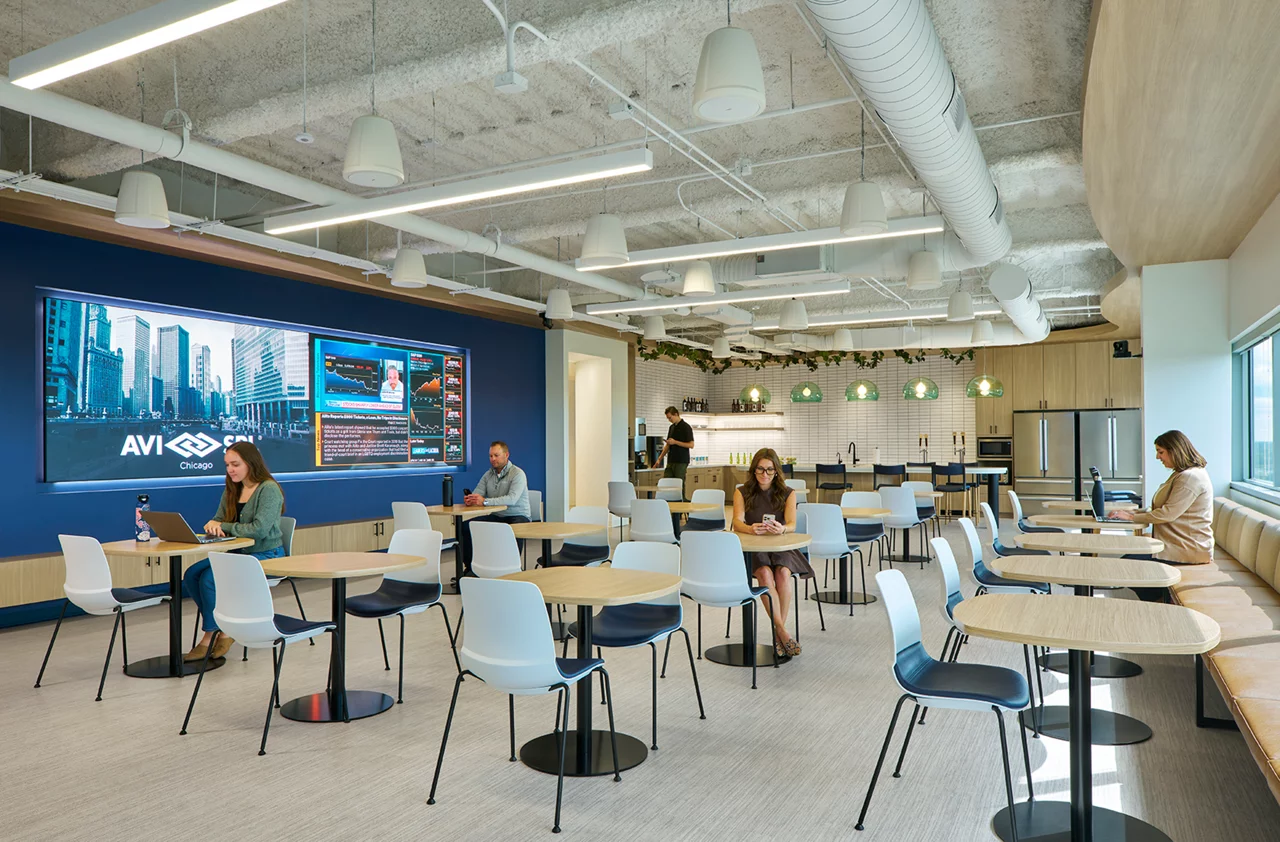 AVI-SPL's modern employee cafe with people working at tables, a large video wall, and a kitchen in the background.