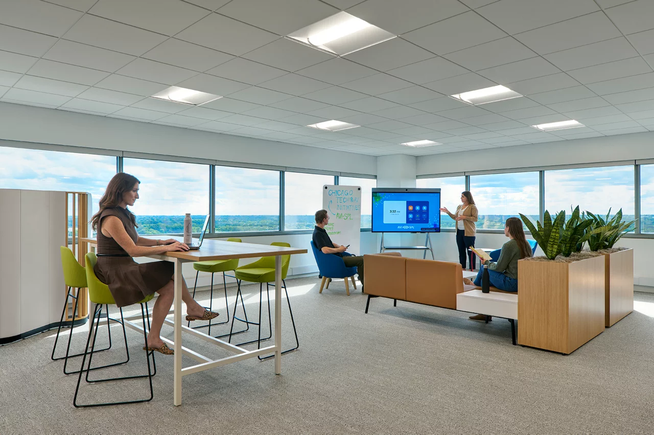 Employees collaborate in a modern AVI-SPL office with flexible furniture, a digital screen, and large, scenic windows.