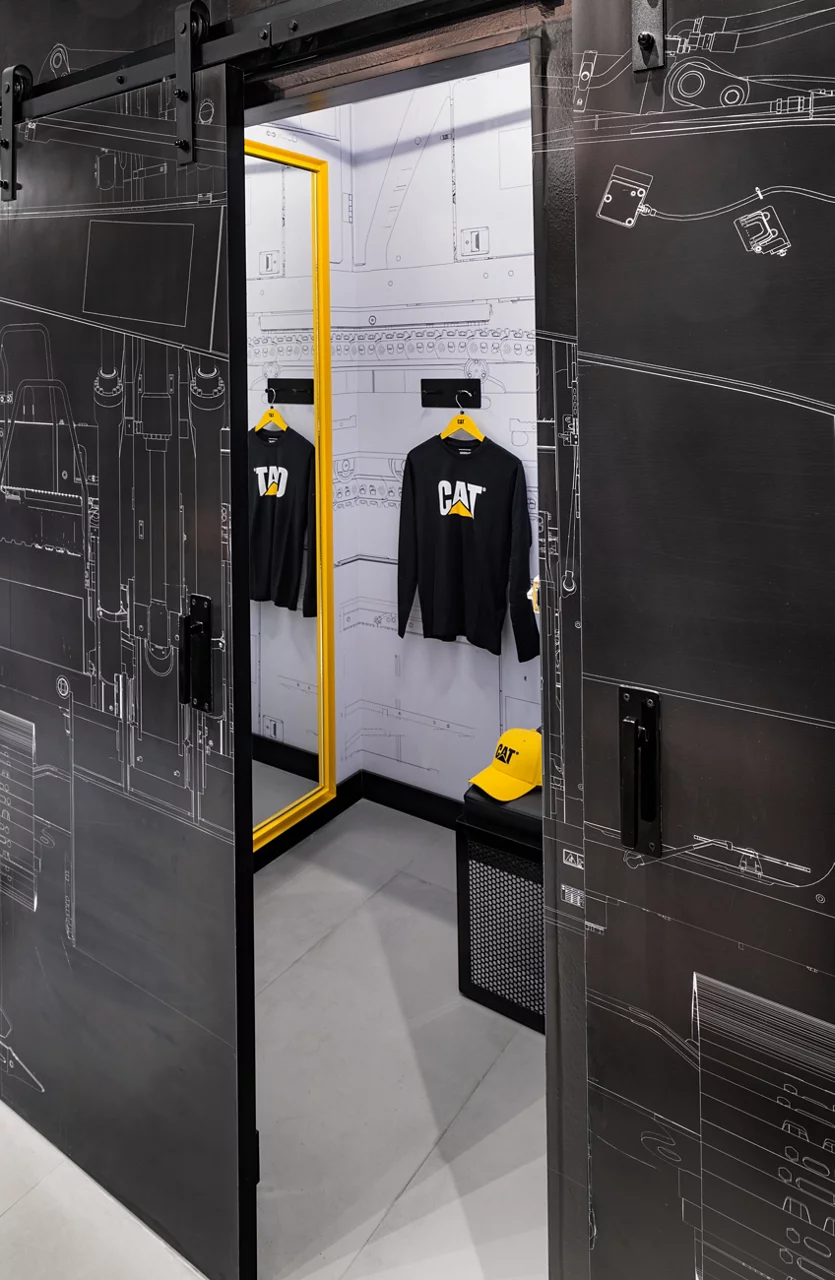 A fitting room in a CAT store with industrial design, featuring machinery blueprint wallpaper and a yellow-framed mirror.