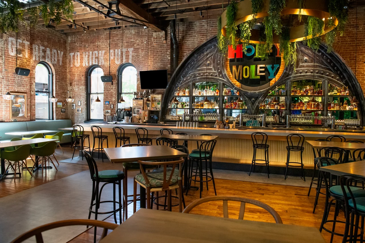 The bar and restaurant at Funlab's Holey Moley, featuring an ornate bar, seating, and a 'Ready to putt loose?' neon sign.