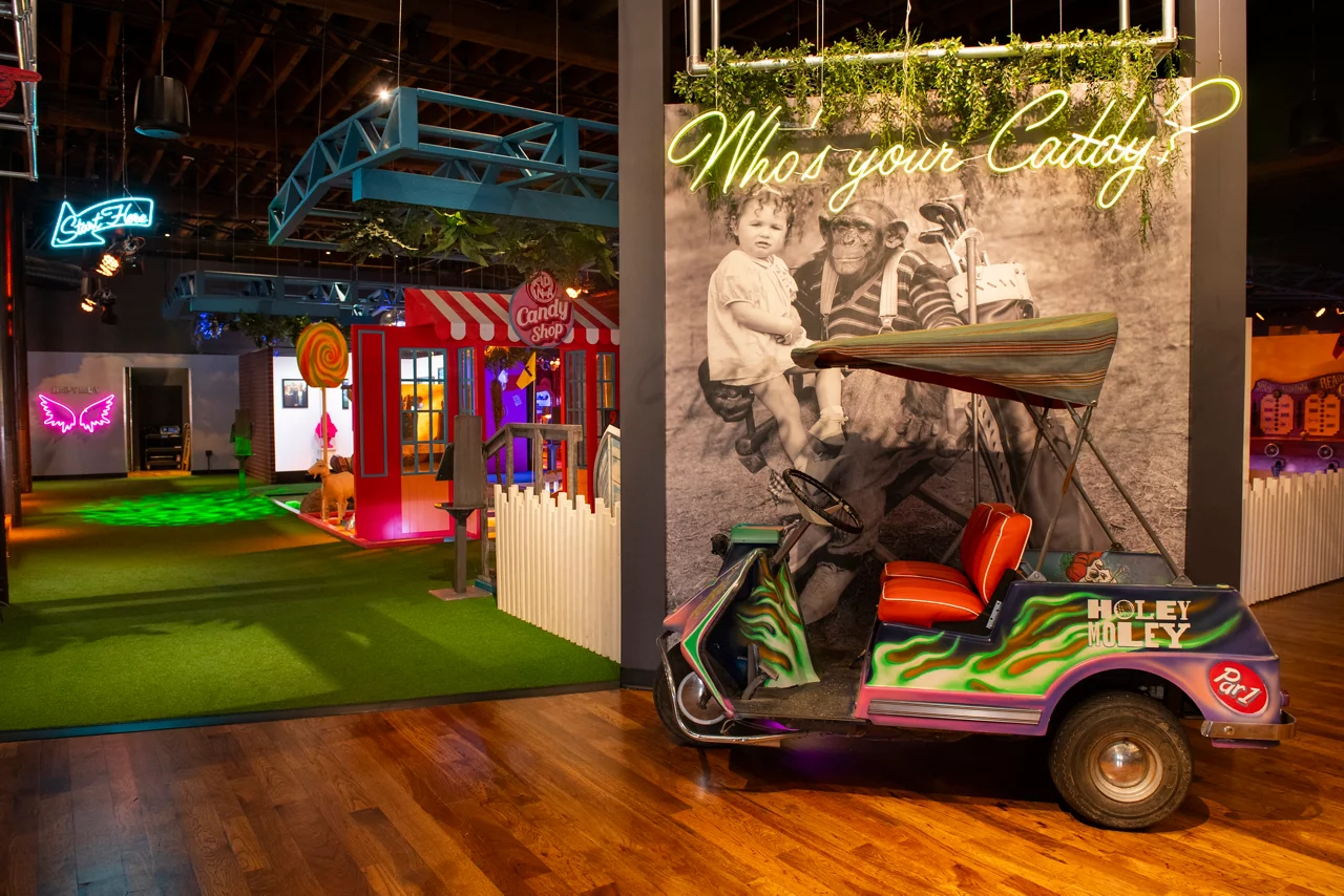 The quirky interior of Funlab's Holey Moley mini-golf, with a custom golf cart and a large 'Who's your Caddy?' mural.