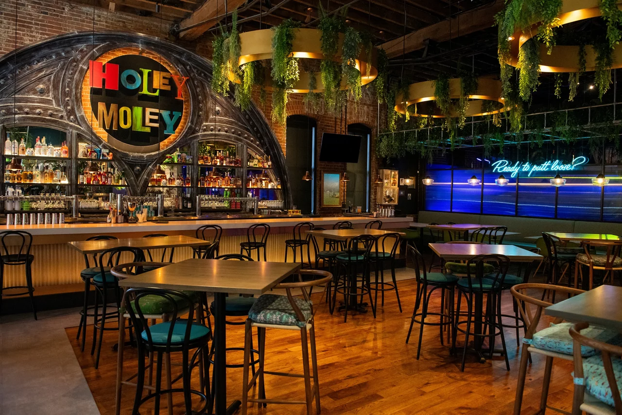 The vibrant bar and seating area at Funlab's Holey Moley venue, featuring an ornate bar, high tables, and hanging greenery.