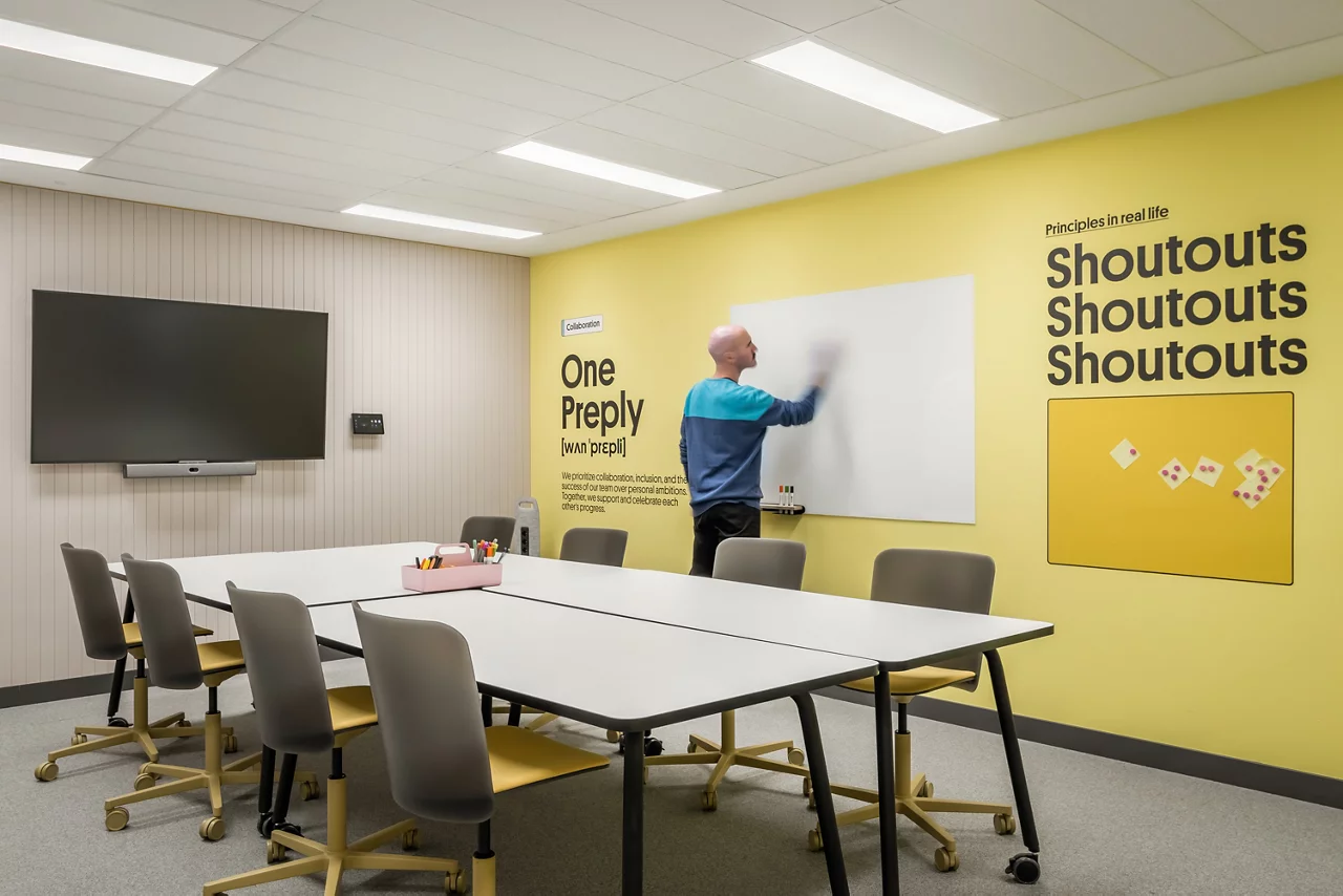A Preply employee uses a whiteboard in a modern meeting room with a yellow wall displaying company values like 'One Preply'.
