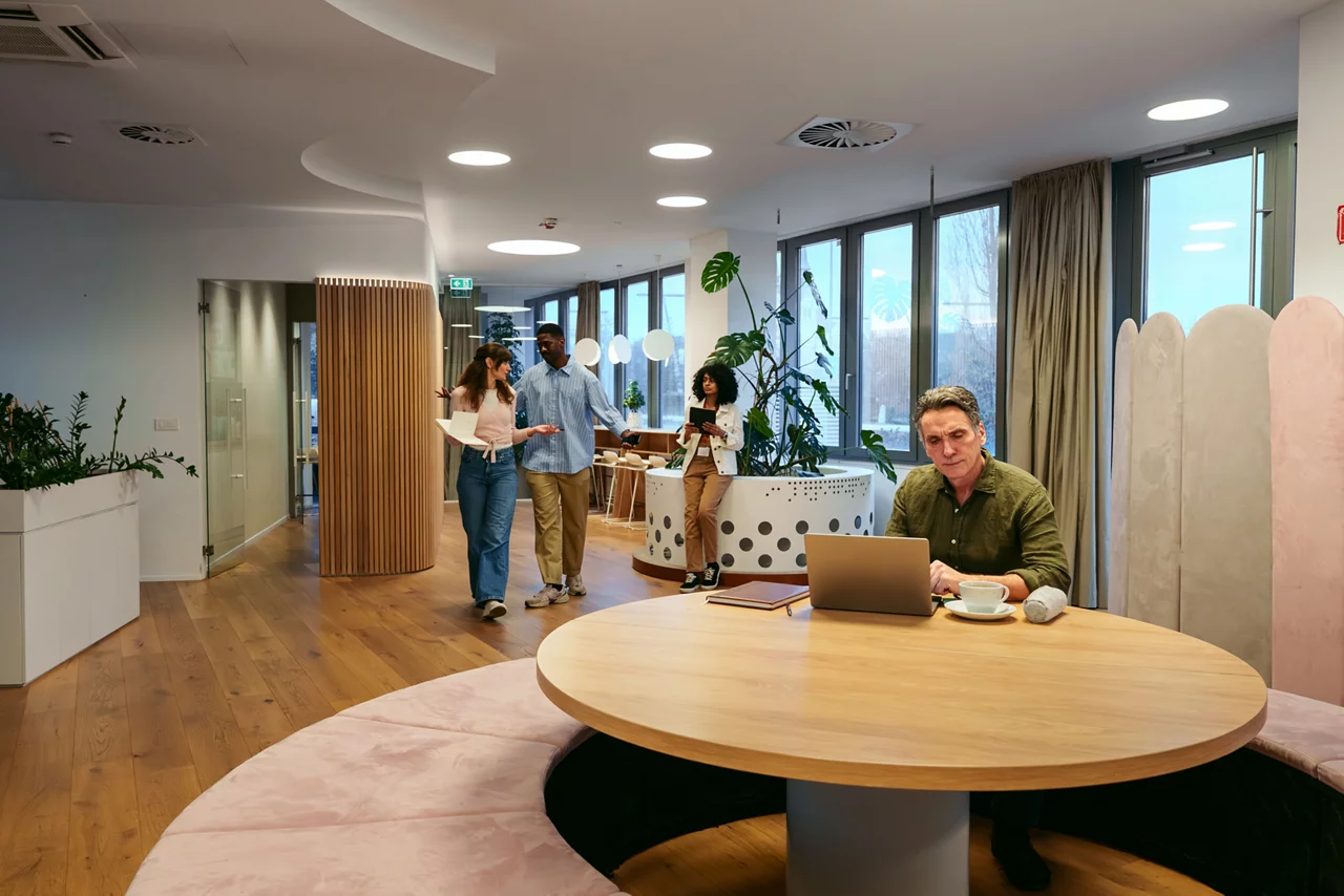 Caucasian businessman focusing on his laptop at a round wooden table in a contemporary open plan office. The space is bright with natural light and features modern decor and plants