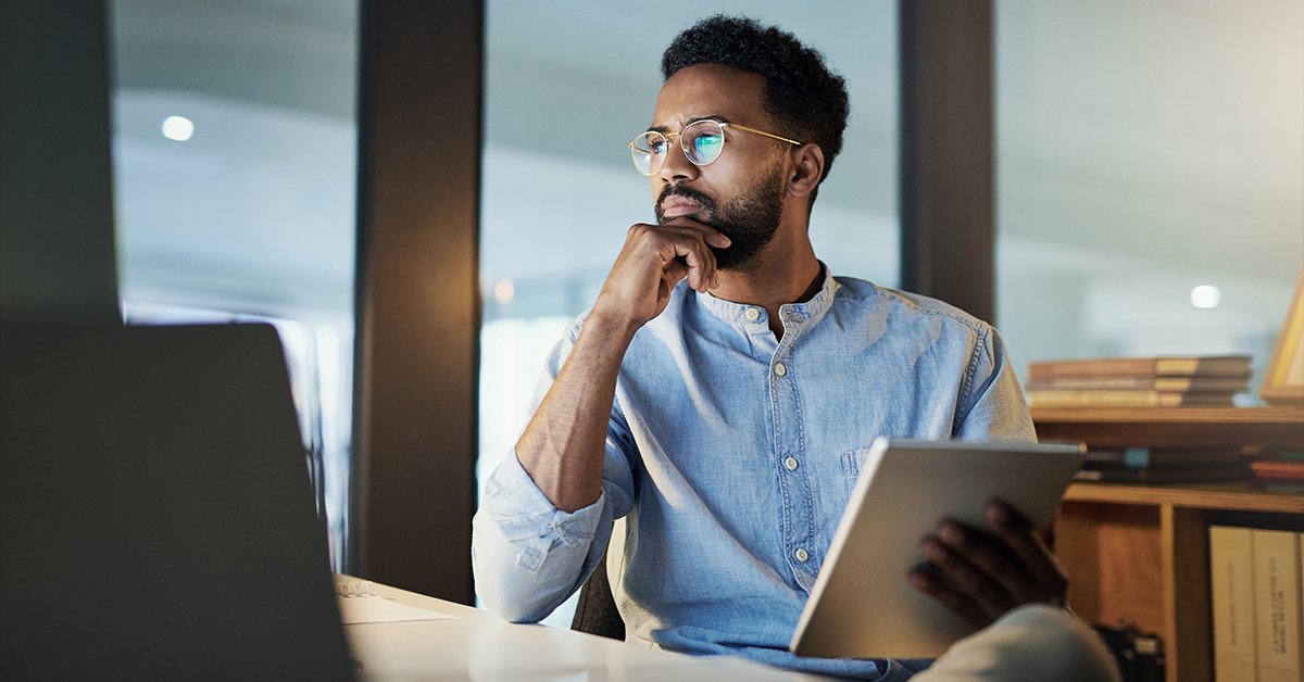 Ein Mann in professioneller Kleidung und mit Brille arbeitet nachdenklich an einem Tablet in einem modernen Büro