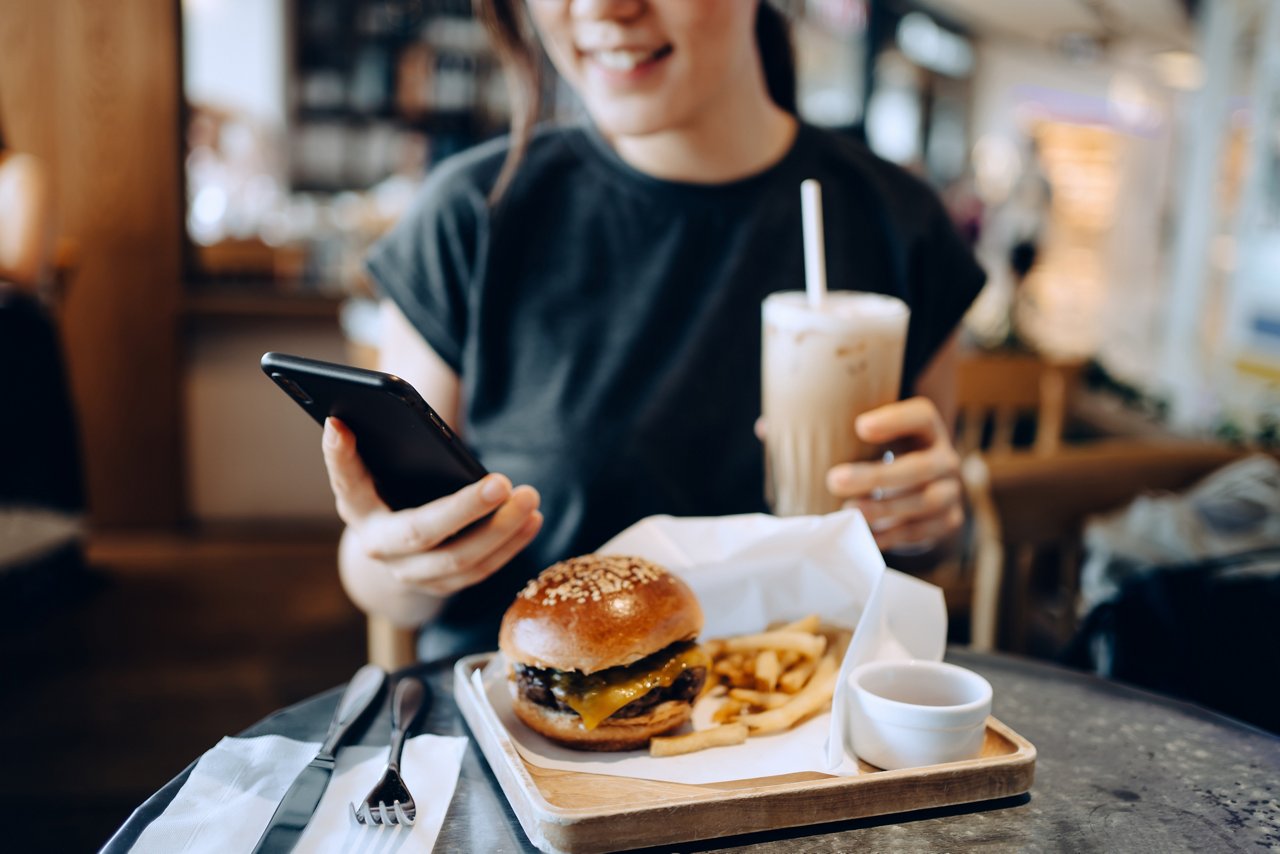 Cropped shot of smiling young Asian woman using smartphone while enjoying her lunch