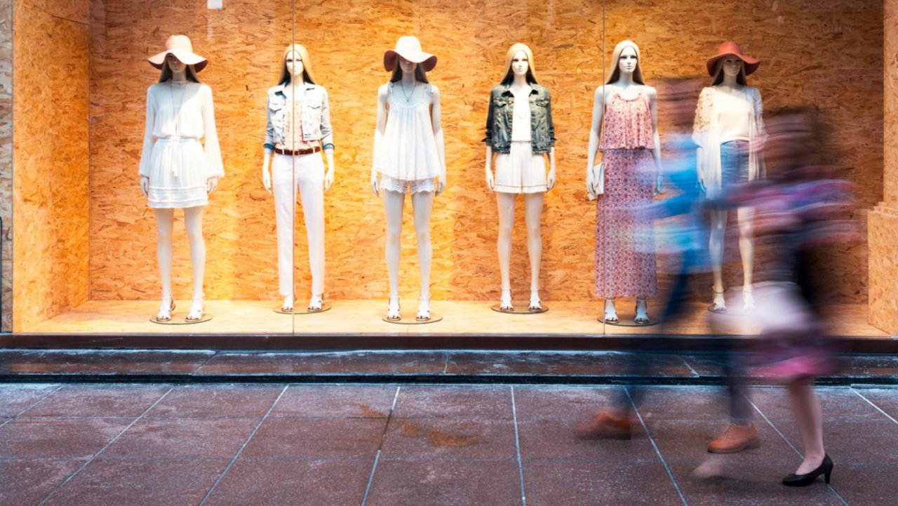 Storefront mannequins in summer outfits with blurred shoppers passing by.