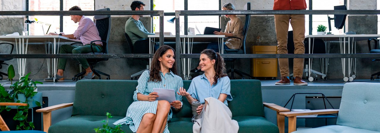 Duas mulheres no centro de uma mesa conversando, com pessoas trabalhando ao fundo.