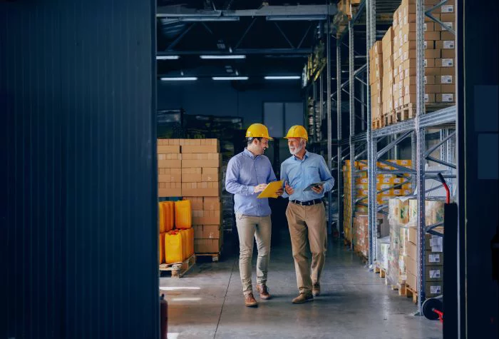 Two warehouse managers wearing yellow hard hats reviewing inventory documents between rows of shelving stacked with boxed products.