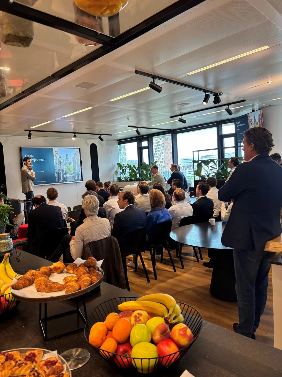 Moderne kantoor vergaderruimte met zakelijke presentatie over retail showcase, deelnemers zitten richting scherm, verfrissingen op de voorgrond.
