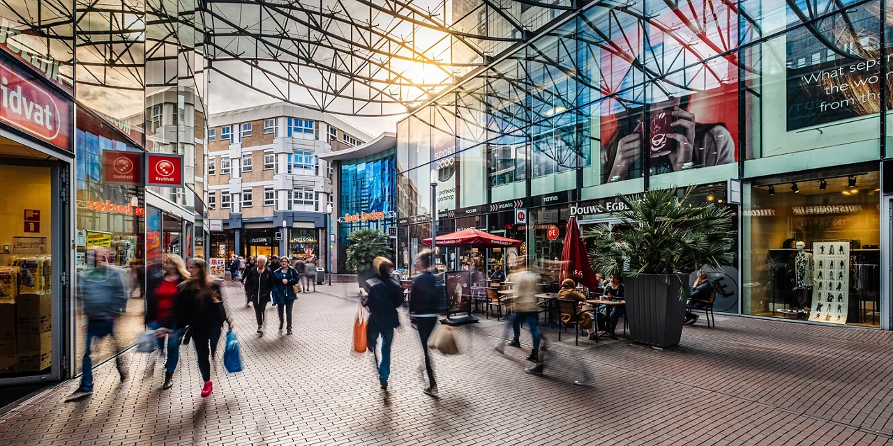 Druk overdekt winkelcentrum met glazen plafond, winkels en zitplaatsen bij cafés terwijl shoppers door de ruimte bewegen.