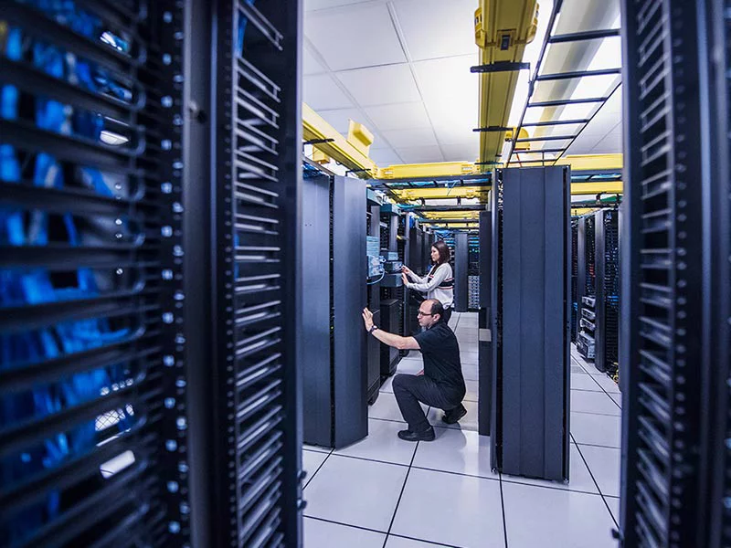 IT technicians working in a modern data center with server racks, blue lighting, and yellow cable trays overhead.