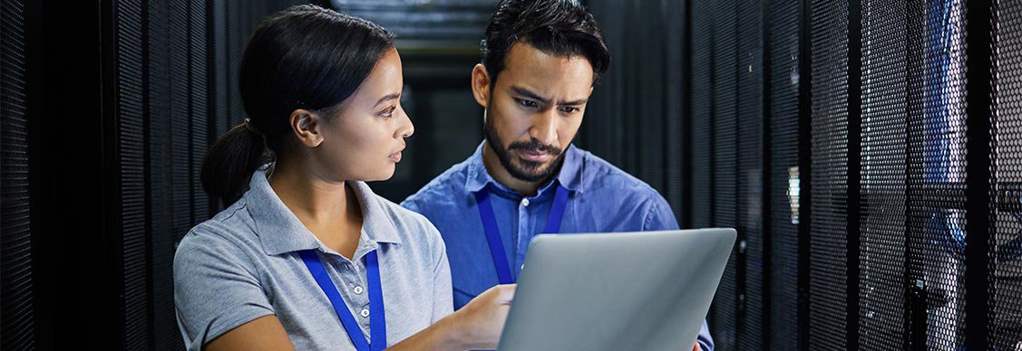 Two individuals in a server room, focused on a laptop, surrounded by rows of servers and technology equipment.