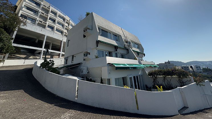 Modern multi-level white building with unique angular architecture featuring terraced balconies and glass elements, surrounded by a curved white wall, set against a blue sky backdrop.