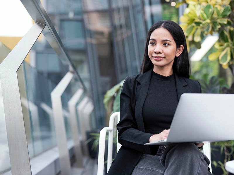 Woman working on laptop outdoors, with greenery and modern building architecture in the background