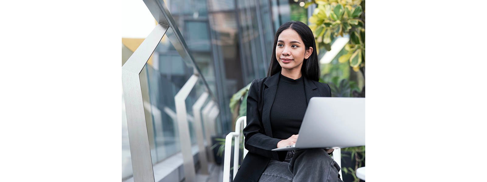 Woman working on laptop outdoors, with greenery and modern building architecture in the background