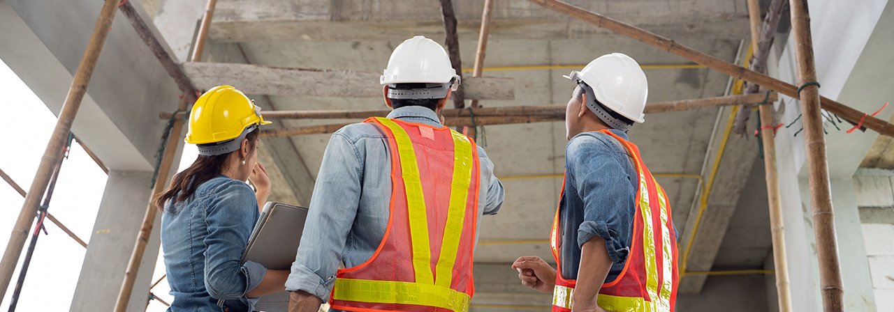 Três profissionais de capacete e colete, de costas, vistoriam uma obra