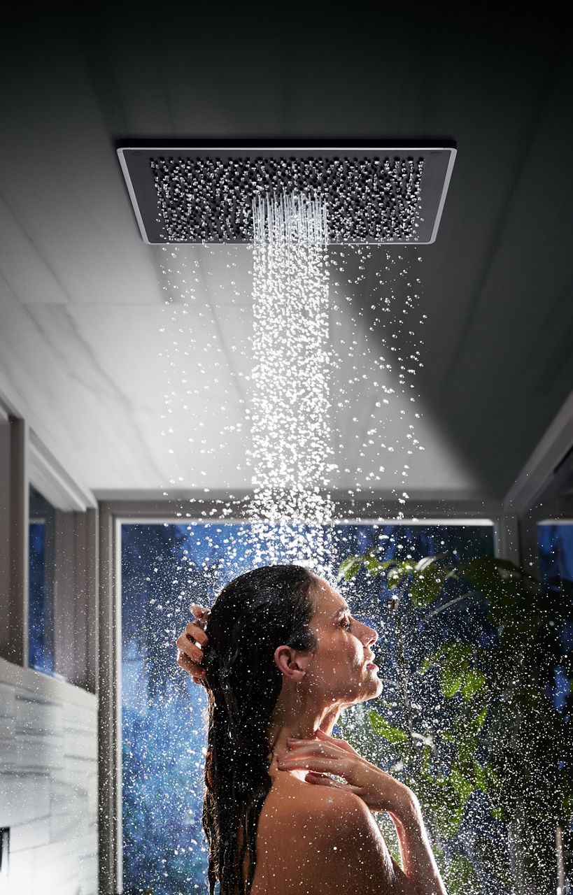 A woman standing under the water from a ceiling mounted Real Rain panel with blue color and green plants in the background.