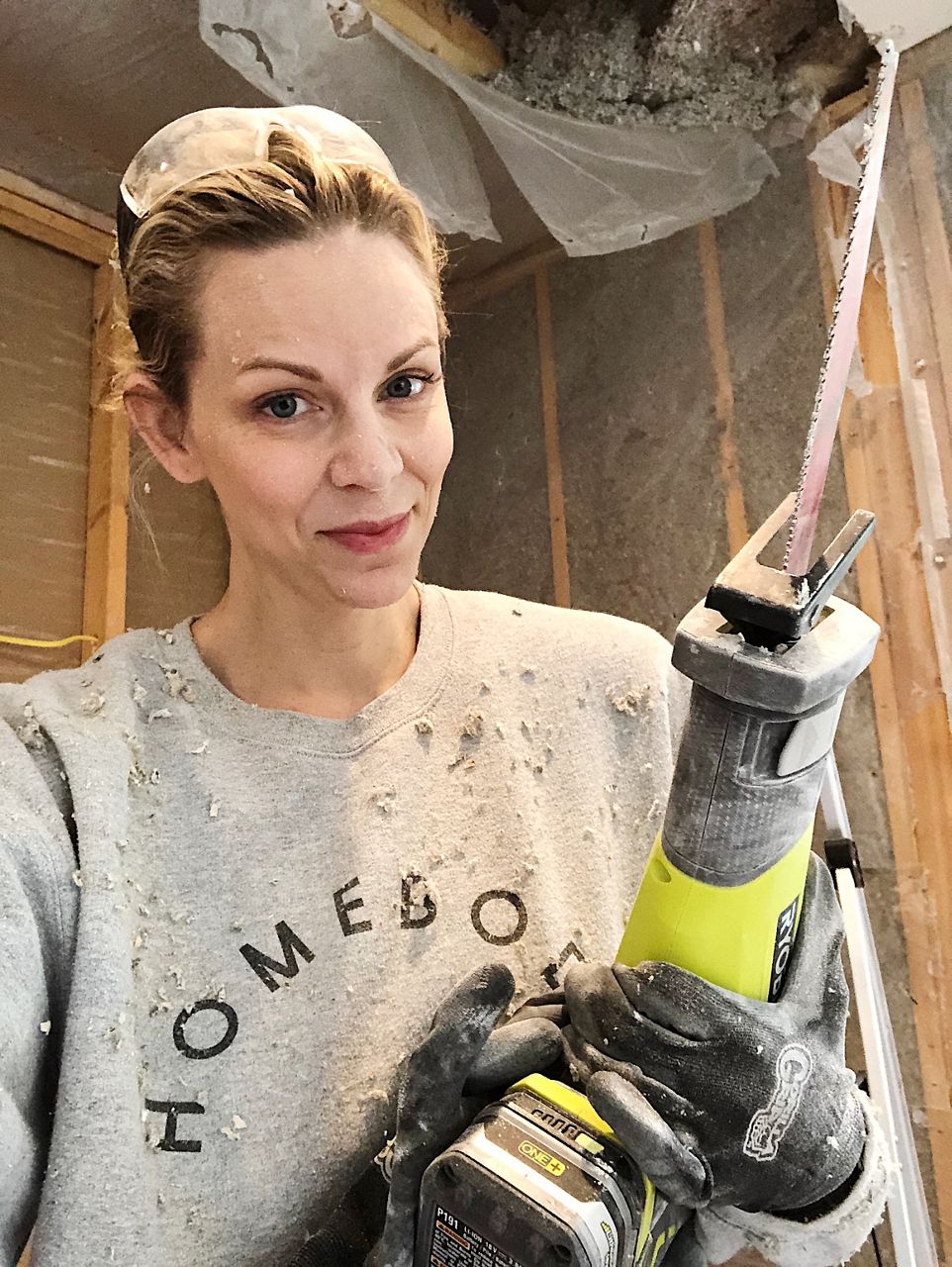 Jennifer Gizzi stands in front of walls stripped to the studs while holding a reciprocating saw.