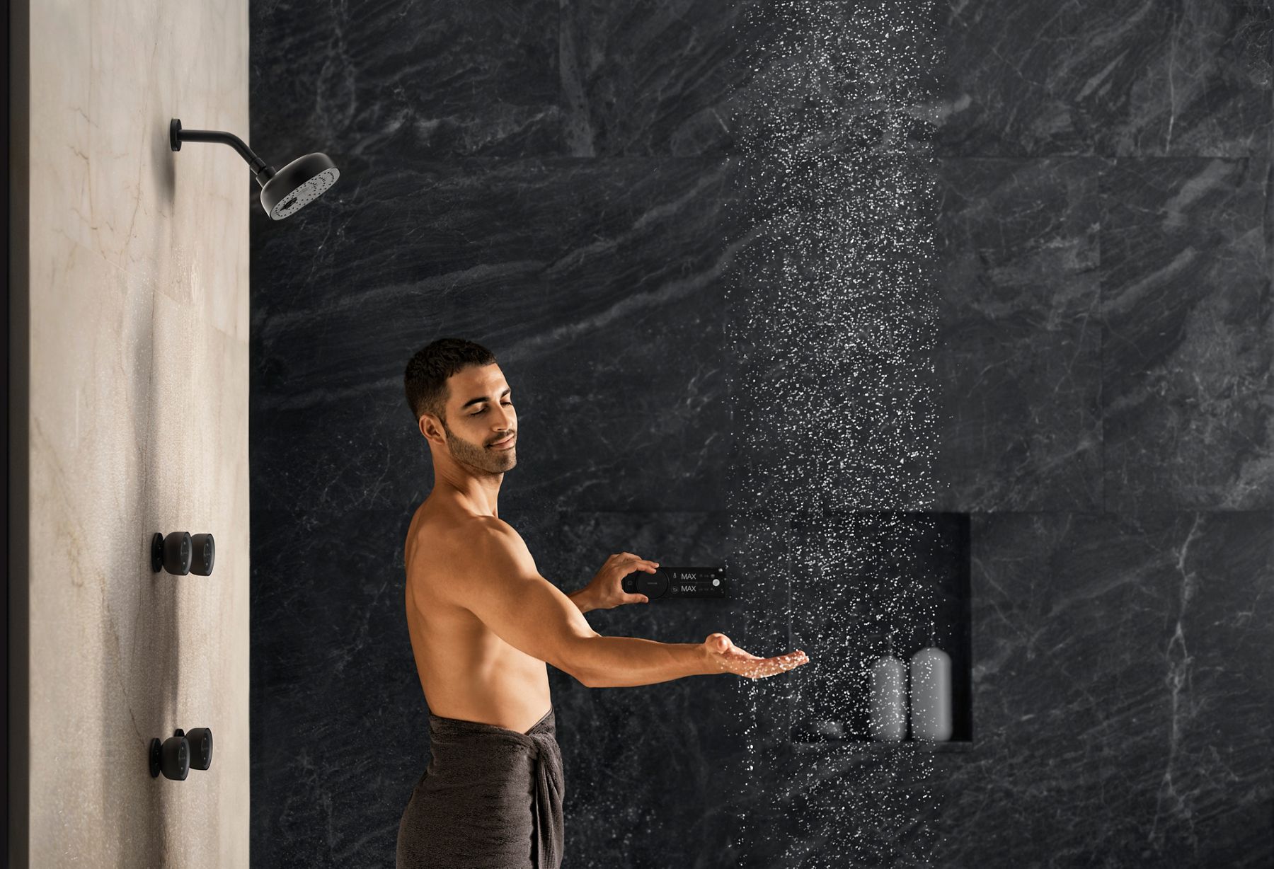 A man wearing a pair of shorts in a large walk-in shower with patterned and striped tile touching the control panel for the digital shower system.