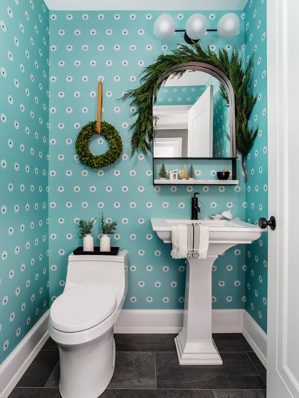 Wide image of a powder room bathroom featuring a fun and funky, polka-dotted teal wall paper, white pedestal sink with matte black, single-lever faucet, one-piece toilet and holiday greenery draped on the mirror and a wreath on the wall. 