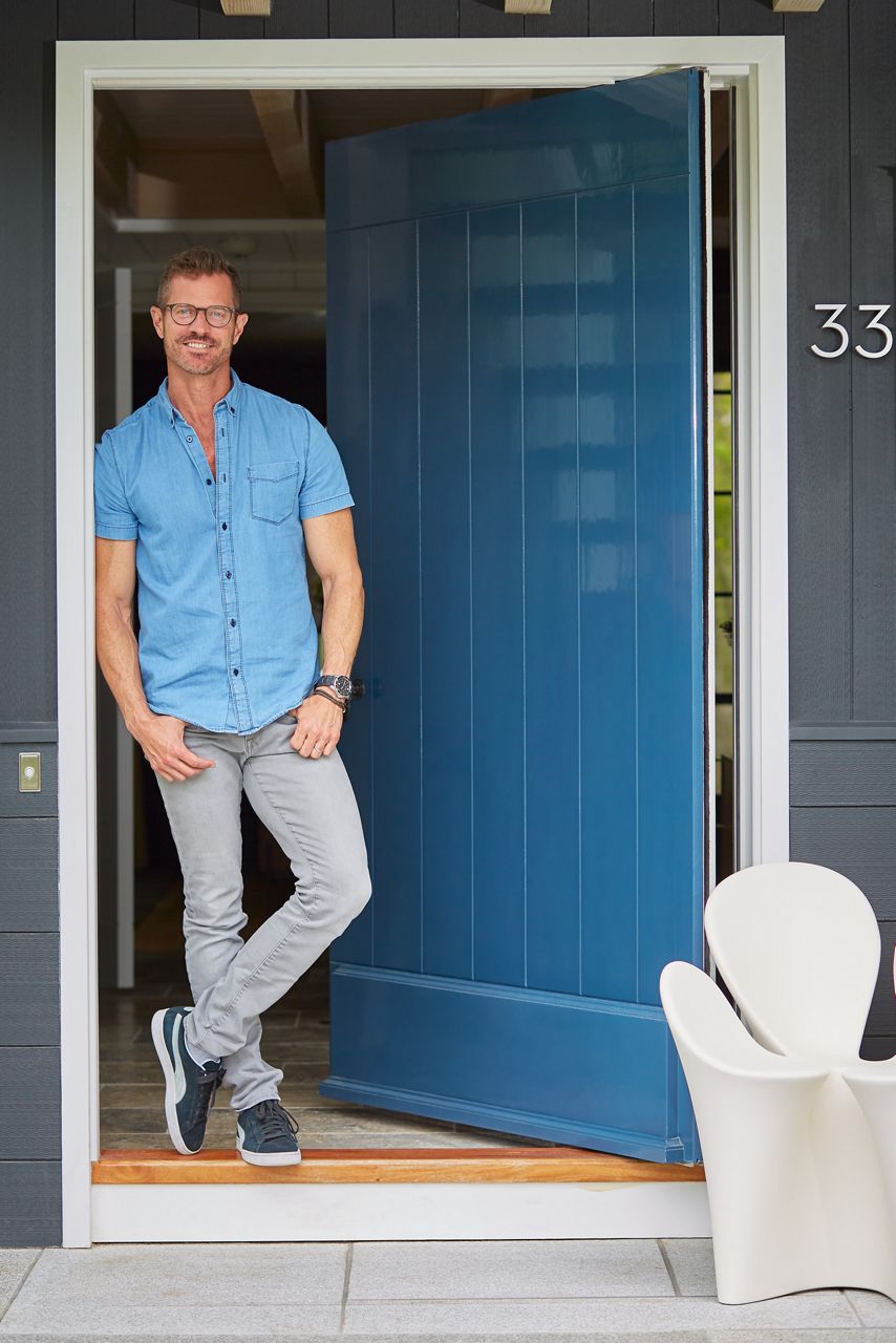 Un Ray Booth souriant, en chemise bleue à manches courtes, en jeans et en chaussures de course, se tient à la porte avant ouverte de sa maison de Provincetown, au Massachusetts.