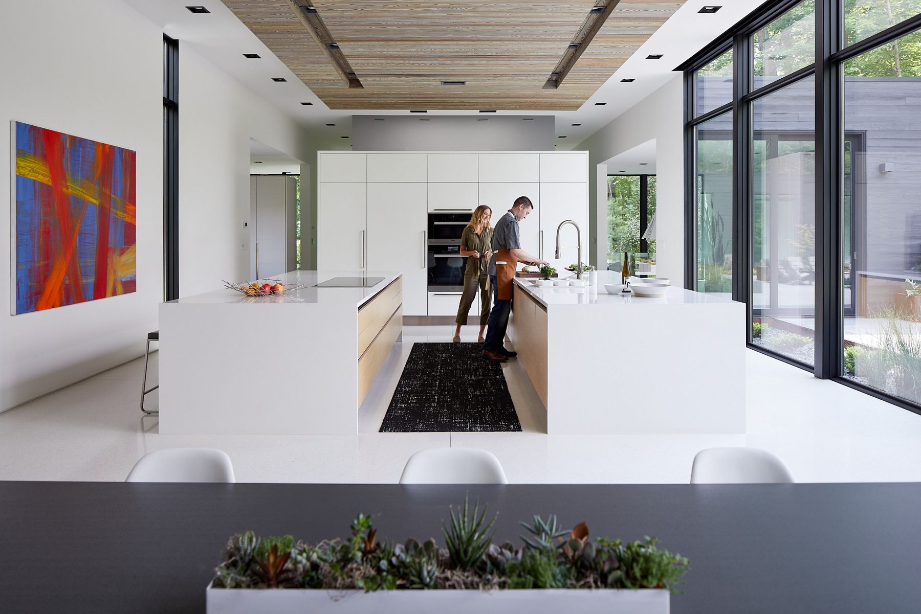 A large white kitchen with two white islands, between which a woman is looking on as a man stands at a sink preparing some green vegetables.