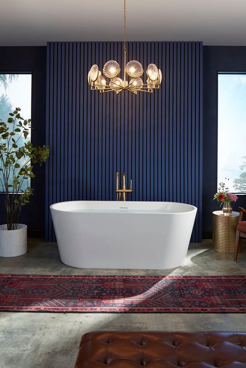 A portrait image shot in daylight displaying a bathroom designed with royal blue wood paneled walls, hard wood flooring, and red Persian rugs. There is a deep tub focused in the center of the shot that is rectangular with rounded sides. The tub faucets are a golden tone, and there is a golden side table to the right of the tub with flowers in a vase and a tall houseplant to the left of the tub. Above the tub is an elegant chandelier that is turned on.​