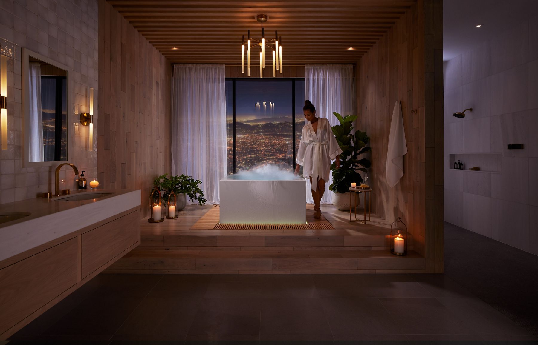 A large bathroom with a woman in the center walking along the side of a Stillness bath, which has steam rising from the surface, with a large window in the background overlooking a nighttime city scape and mountains in the distance.