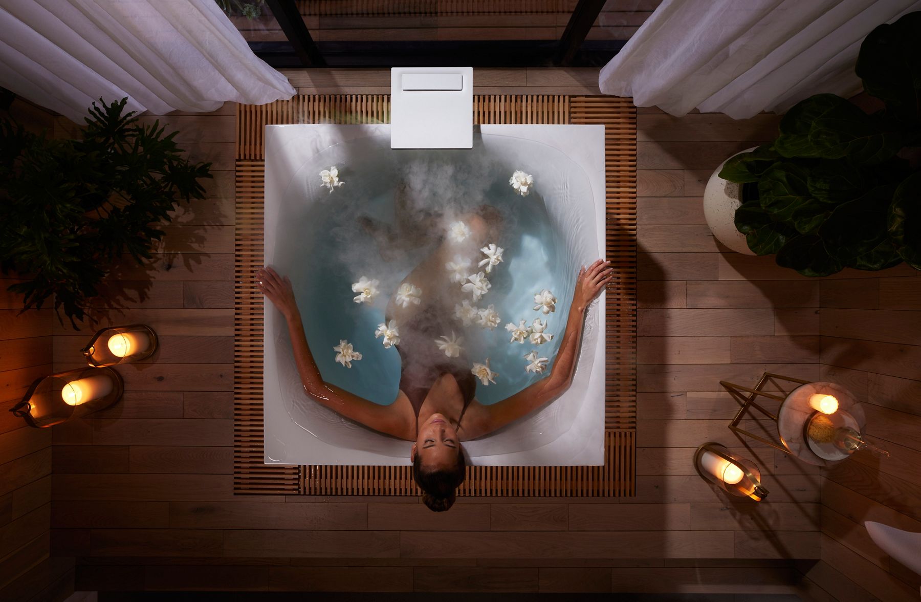Overhead view of a woman soaking in a luxury bathtub, surrounded by candles and flowers.