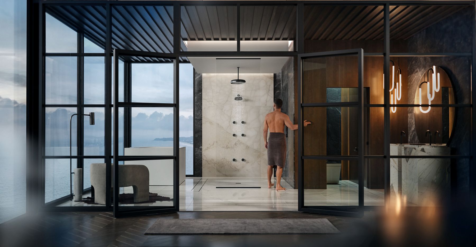 A man in a towel standing in a large glass walled bathroom with a shower area in the center, a freestanding bath on the left, and a vanity area and a toilet in an alcove on the right.