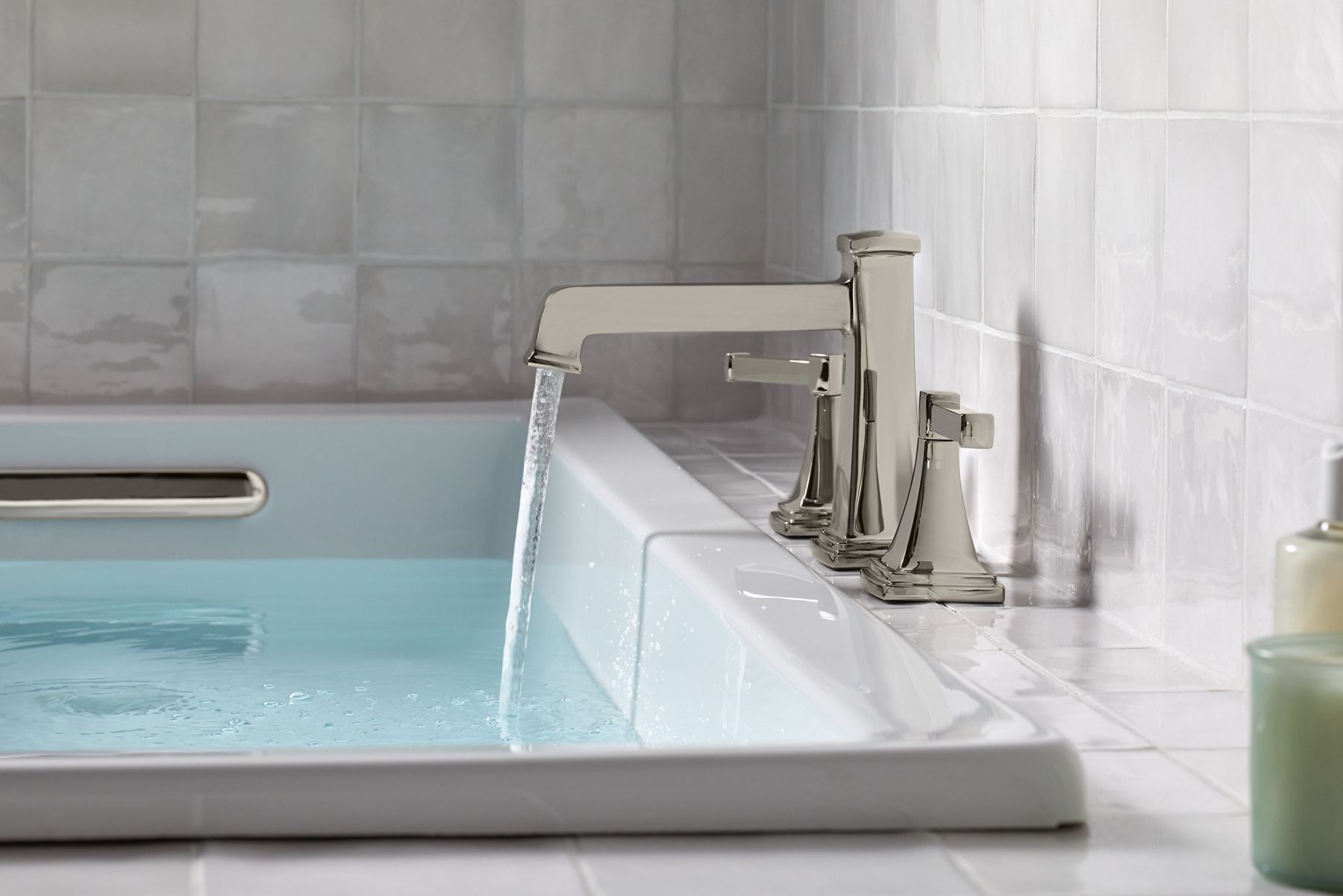 Water flows from a deck-mounted KOHLER Riff bath spout with lever handles into a white rectangle bath with white tiled walls and surround.