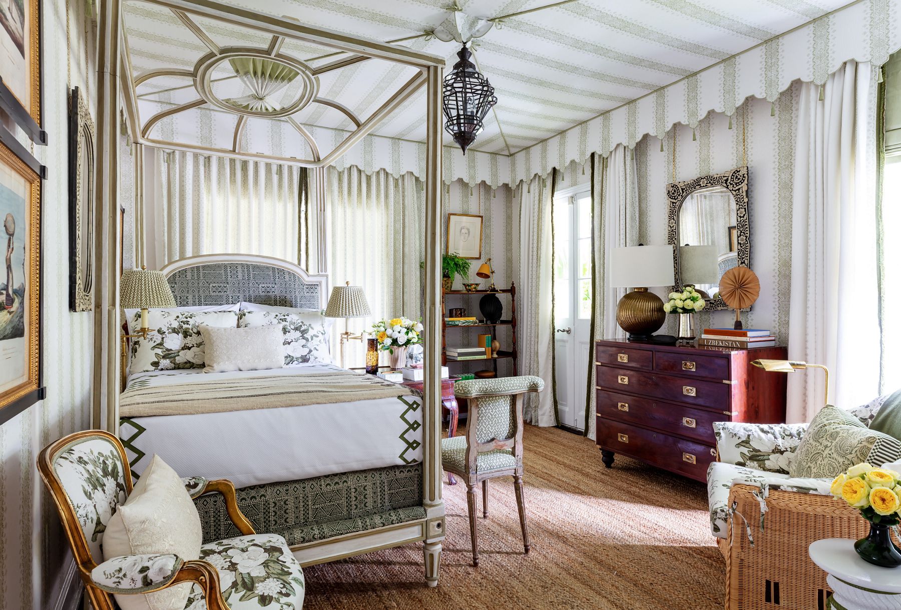 A Grandmillennial style bedroom with green and white striped wallpaper, canopy bed, bamboo display shelf, and an assortment of chairs in contrasting styles and upholstery patterns.