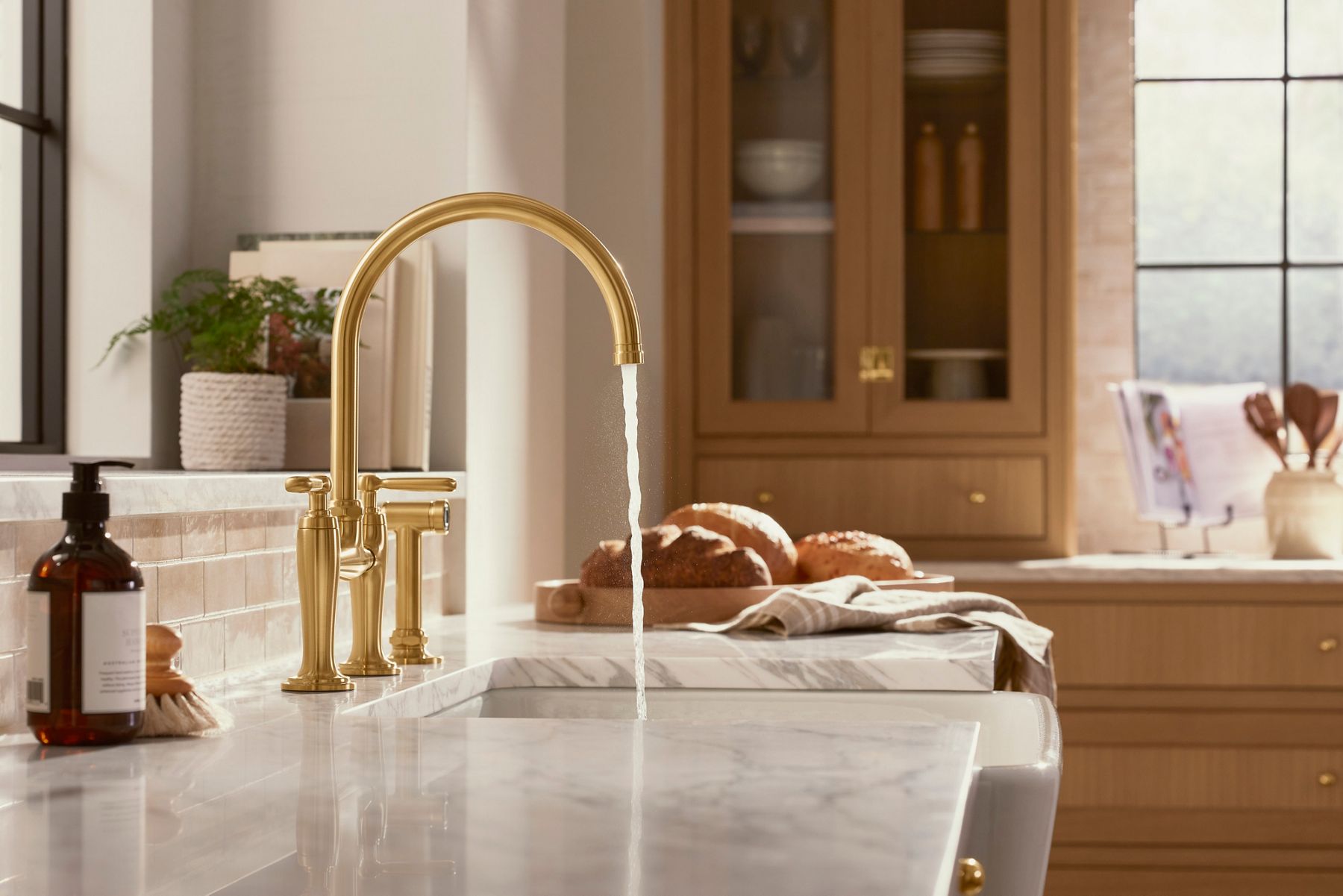Elegant kitchen with a white marble countertop featuring a gold faucet. Water flows from the faucet into the sink. In the background, there are wooden cabinets, fresh bread on a cutting board, and kitchen decor, including a potted plant and a bottle of soap on the counter.