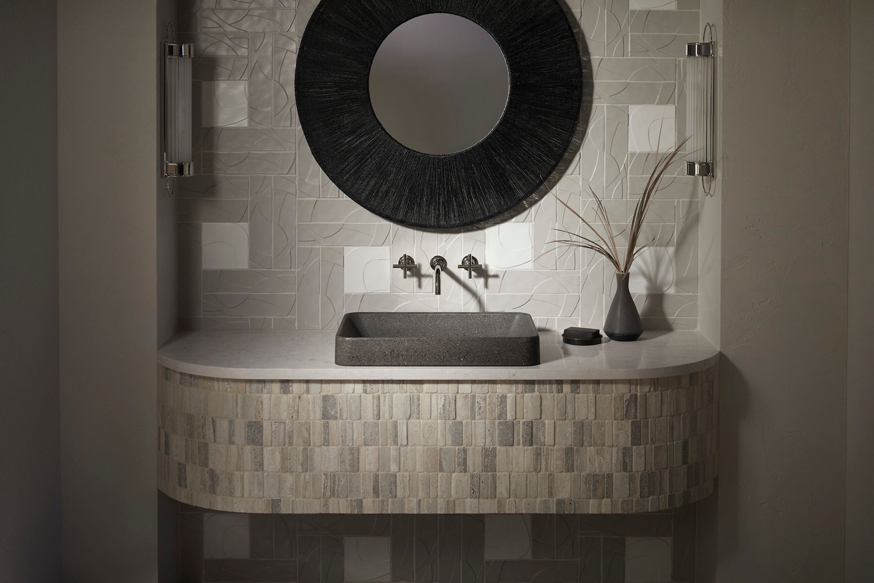 A bathroom styling space with a gray rectangular sink made from recycled material, a back wall of light gray tile with surface layers, a tiled vanity counter, and a round mirror with a wide black frame.