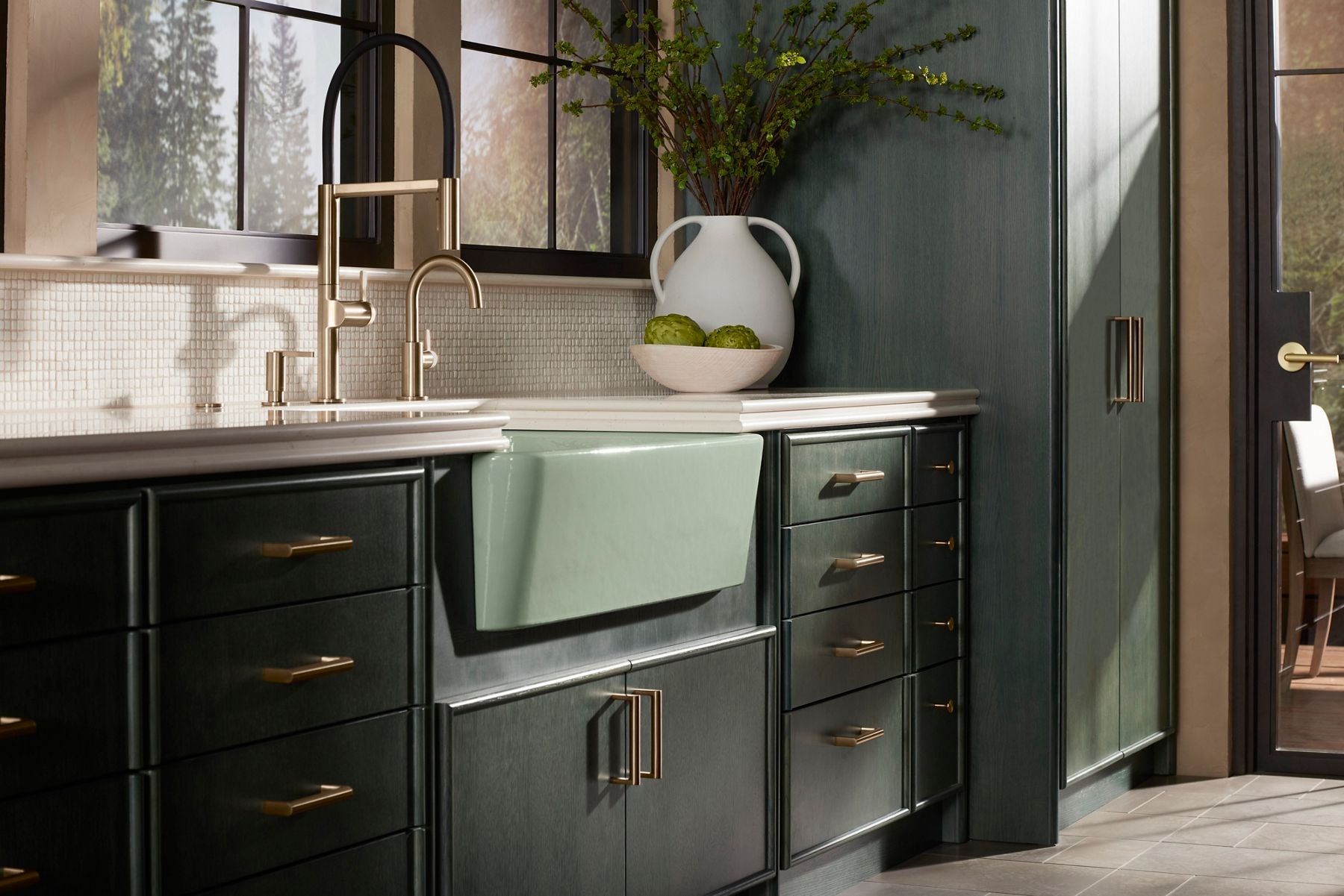 Light green apron-front farmhouse sink with a brass bridge faucet in a dark cabinetry kitchen.