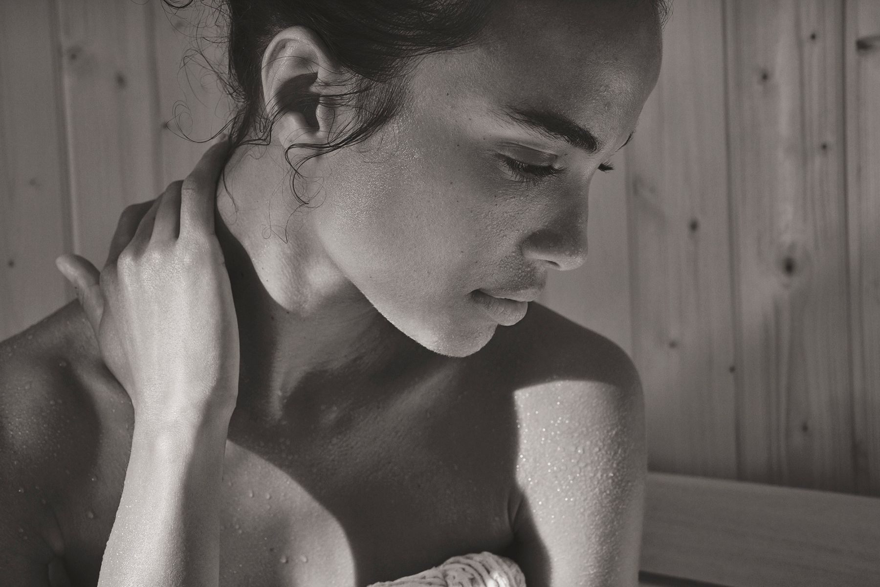 A close-up of a woman wrapped in a towel sitting in a sauna, gently touching her neck.