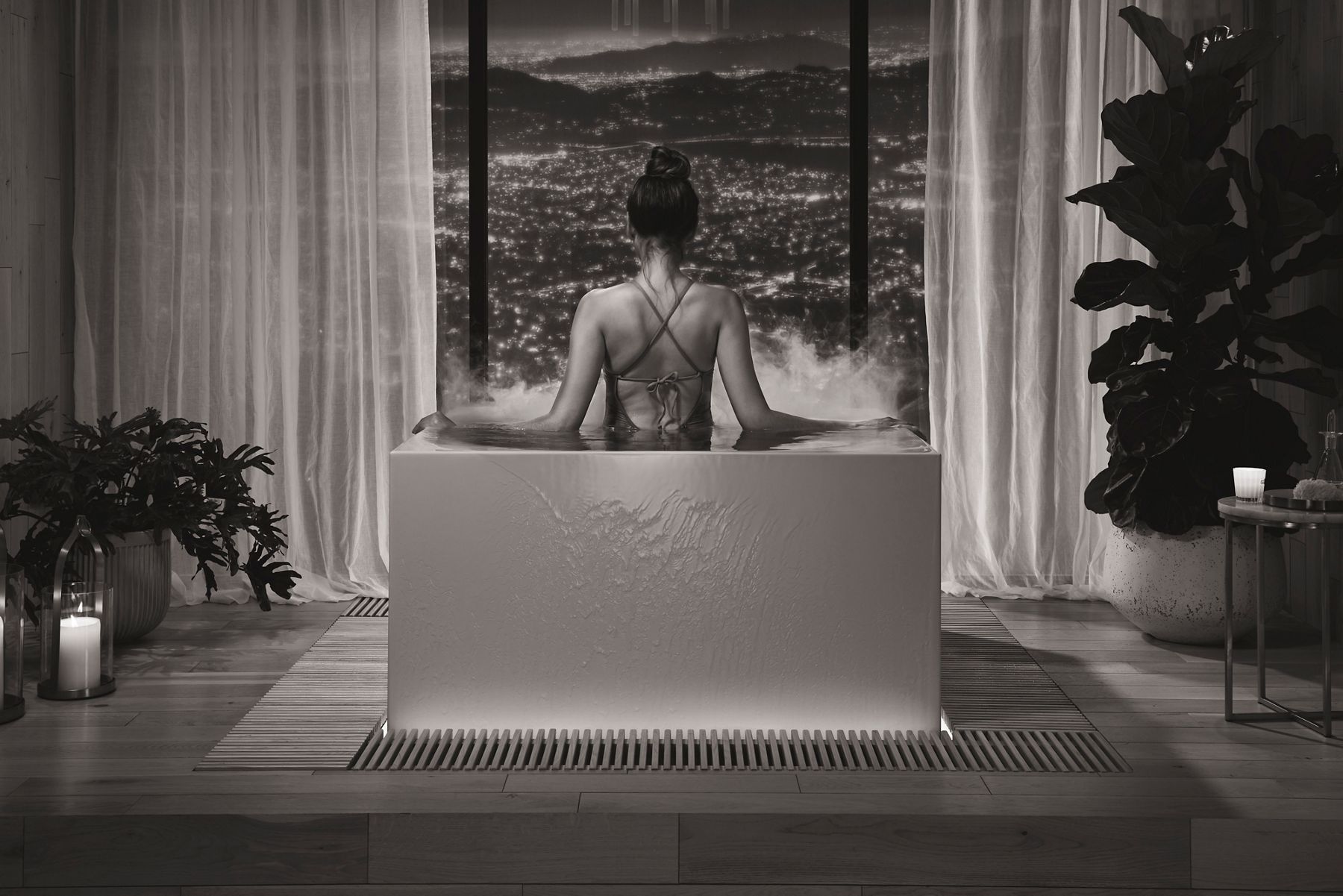 Woman sitting in a bathtub facing large windows with a scenic city view, surrounded by candles and plants.