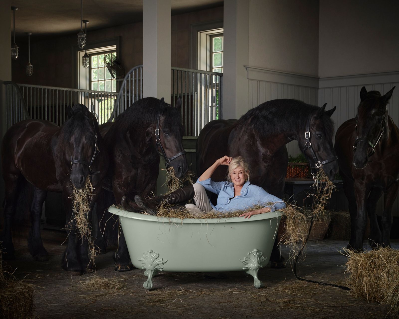 Martha Stewart lounging in a vintage clawfoot bathtub filled with hay, surrounded by three black horses in a barn setting.