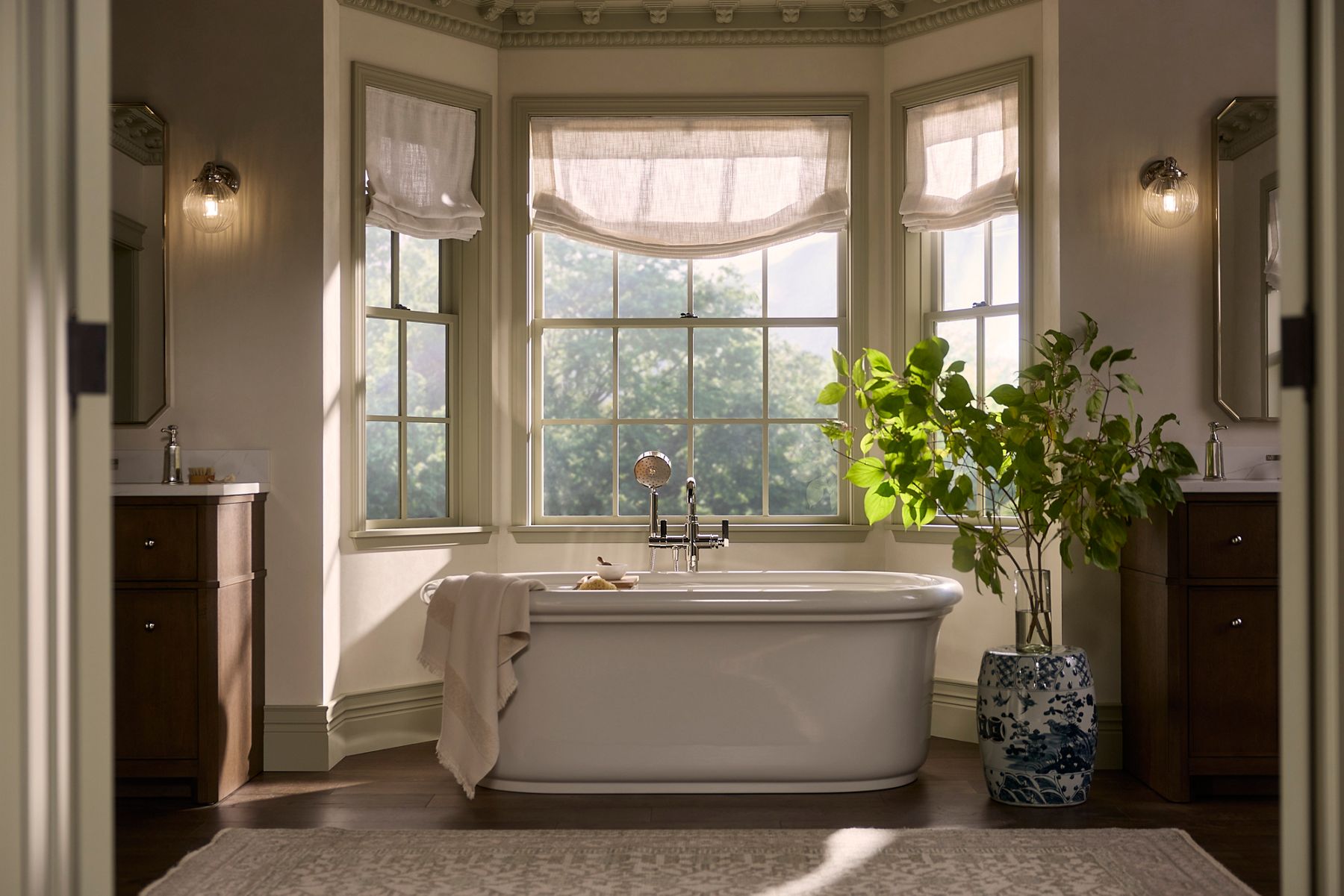 Warm, elegant bathroom with KOHLER Claude freestanding bath by a window with natural afternoon light, ceramic pottery, greenery, and dark wood vanities.