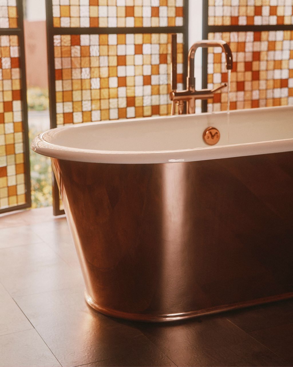 A copper freestanding enameled cast iron bath in the Flamingo Estate Bathhouse by Kohler, surrounded by stained glass panels in warm earth tones.